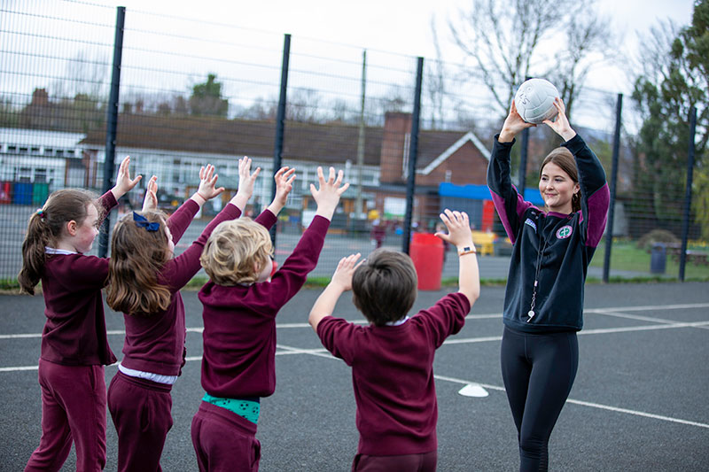 Female student coaching basketball with primary school children